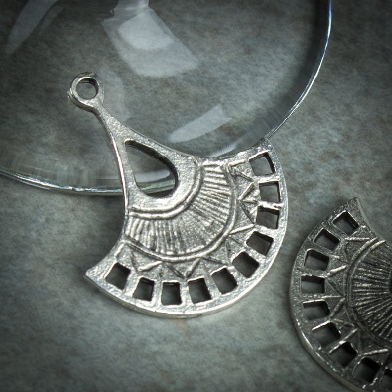 Puede incluir: Dos colgantes de metal plateado en forma de abanico con un dise&ntilde;o geom&eacute;trico. Los colgantes tienen un agujero en la parte superior para sujetarlos a un collar o pulsera.
