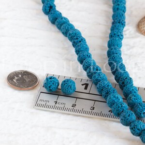 May include: Two teal blue lava rock beads measuring 1 cm in diameter, next to a ruler. A strand of teal blue lava rock beads is in the background.