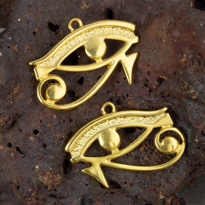 May include: Two gold-colored metal charms in the shape of the Eye of Horus, an ancient Egyptian symbol of protection. The charms are detailed with intricate lines and a raised circle in the center of each eye.