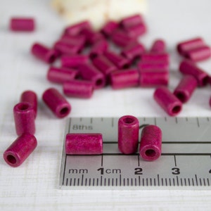 Puede incluir: Primer plano de una regla con una cuenta de madera rosa junto a la marca de 1 cm. La cuenta mide aproximadamente 1 cm de largo. Hay muchas otras cuentas rosas esparcidas en el fondo.