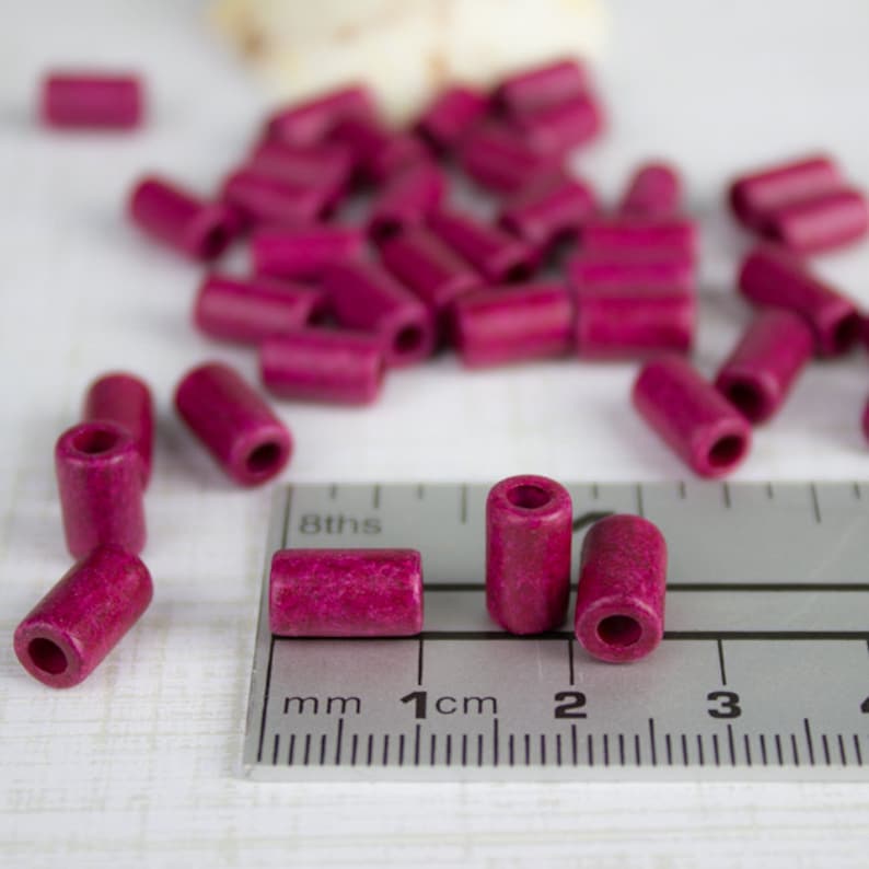 Puede incluir: Primer plano de una regla con una cuenta de madera rosa junto a la marca de 1 cm. La cuenta mide aproximadamente 1 cm de largo. Hay muchas otras cuentas rosas esparcidas en el fondo.