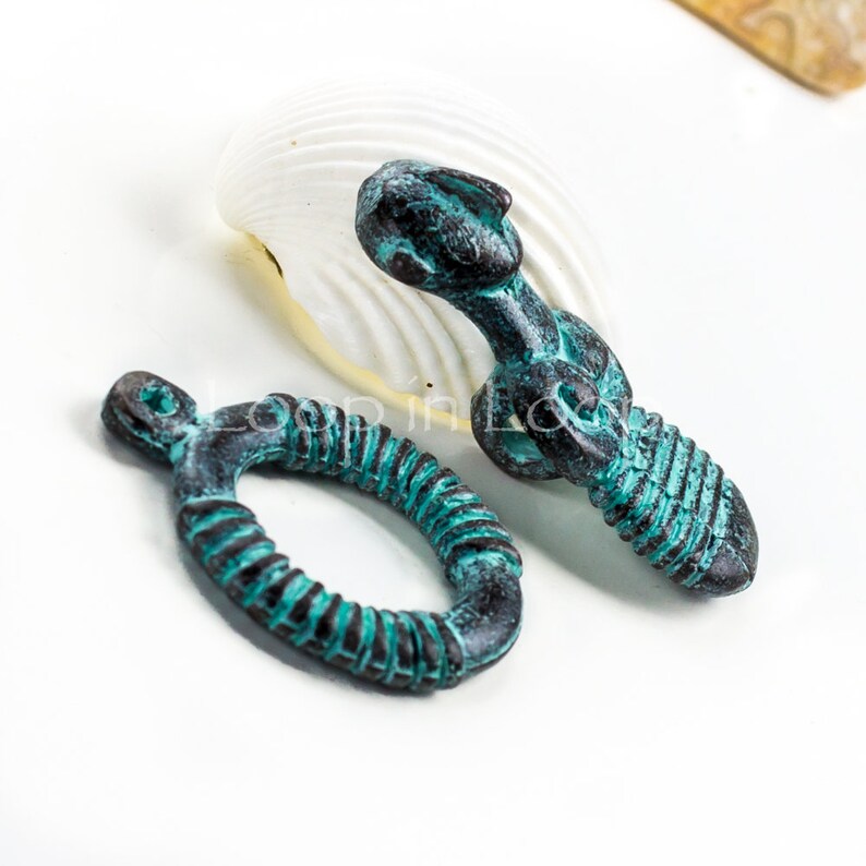 May include: Two teal green metal jewelry findings. One is a ring with a twisted design and the other is a small, detailed figure.