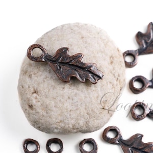 May include: A close-up of several antique copper-toned oak leaf charms with a hole for stringing. The charms are on a white surface.