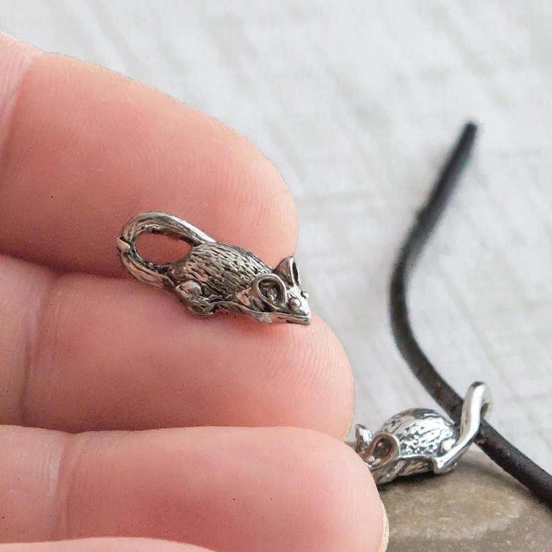 Puede incluir: Un dije de rat&oacute;n plateado con textura de pelaje detallada y orejas grandes. El dije tiene un lazo para sujetarlo a un collar o pulsera. El rat&oacute;n se muestra con un cord&oacute;n negro.