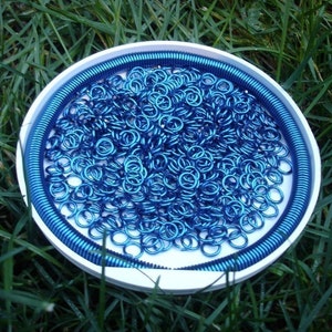 May include: A white circular container filled with a large quantity of small, metallic blue jump rings. A continuous blue coiled wire lines the inner perimeter of the container, creating a border around the loose collection of circular metal rings.