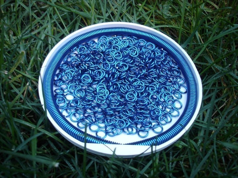 May include: A white circular container filled with a large quantity of small, metallic blue jump rings. A continuous blue coiled wire lines the inner perimeter of the container, creating a border around the loose collection of circular metal rings.