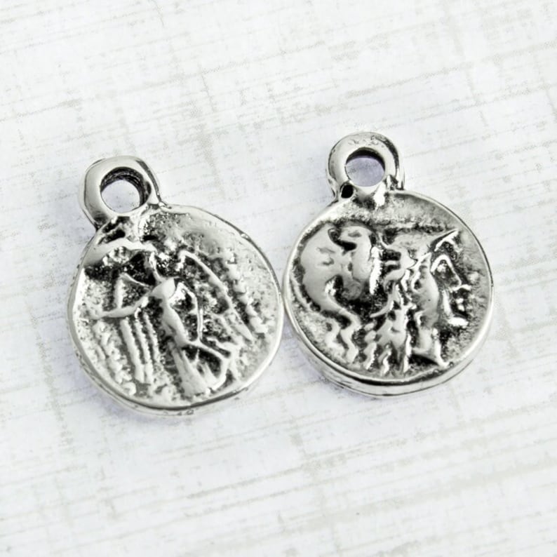 May include: Two silver-toned metal charms with a circular design. One charm features a winged figure, while the other charm features a profile of a person with a winged figure.