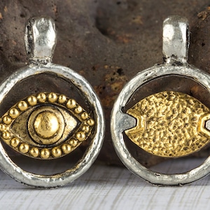 May include: Two silver charm pendants with a gold eye design. The eye on the left pendant is surrounded by small gold beads. The eye on the right pendant is textured.
