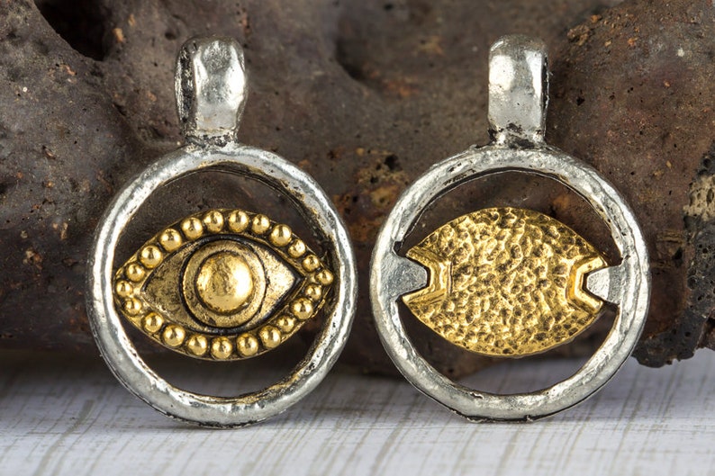 May include: Two silver charm pendants with a gold eye design. The eye on the left pendant is surrounded by small gold beads. The eye on the right pendant is textured.