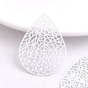 May include: Two white metal leaf-shaped earrings with a delicate, intricate design. The earrings are cut out and have a teardrop shape.