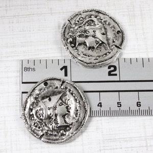 K&ouml;nnte beinhalten: Zwei silberfarbene Metallm&uuml;nzen-Anh&auml;nger mit erhabenen Designs. Die Anh&auml;nger haben einen Durchmesser von etwa 2,5 cm und zeigen auf der einen Seite ein Profil eines Frauenkopfes und auf der anderen Seite ein Design eines Frauenkopfes mit einer Krone.