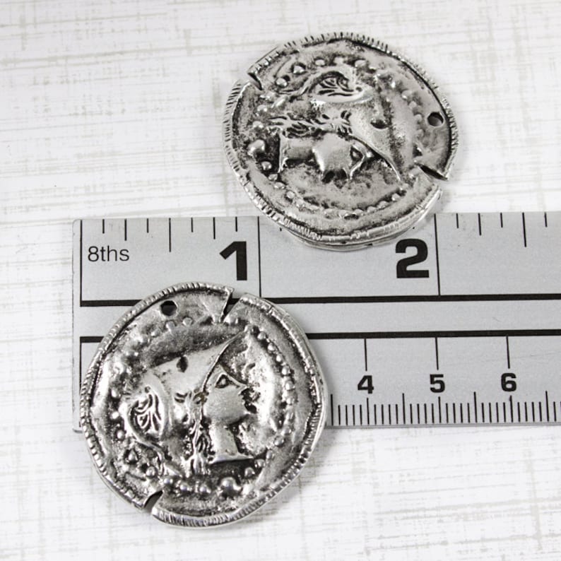 K&ouml;nnte beinhalten: Zwei silberfarbene Metallm&uuml;nzen-Anh&auml;nger mit erhabenen Designs. Die Anh&auml;nger haben einen Durchmesser von etwa 2,5 cm und zeigen auf der einen Seite ein Profil eines Frauenkopfes und auf der anderen Seite ein Design eines Frauenkopfes mit einer Krone.