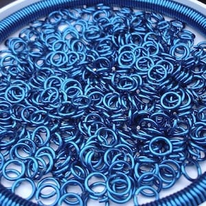 May include: A collection of small, metallic blue jump rings piled together inside a circular white container. The container features a ribbed blue border along the inner edge, framing the loose, circular metal components in a vibrant blue hue.