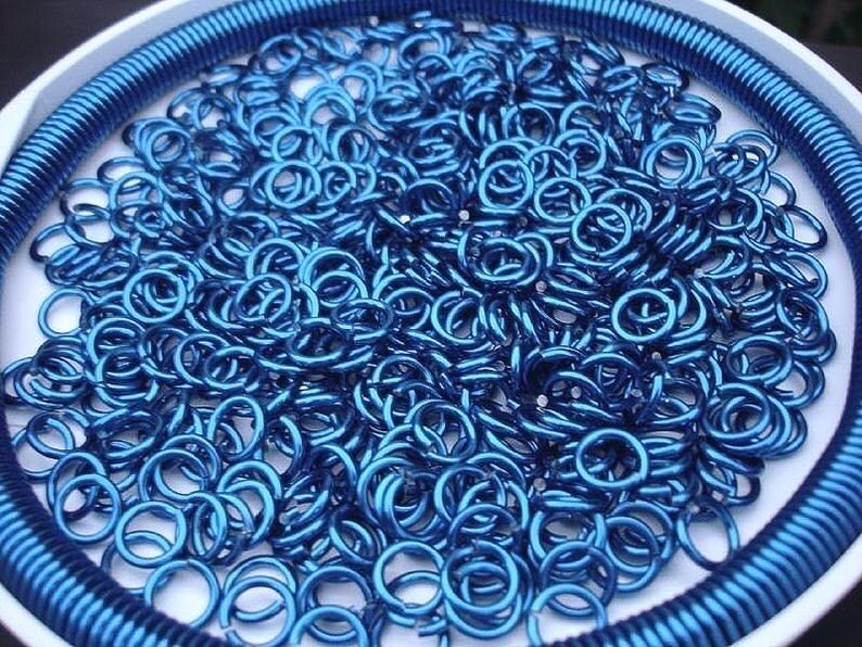 May include: A collection of small, metallic blue jump rings piled together inside a circular white container. The container features a ribbed blue border along the inner edge, framing the loose, circular metal components in a vibrant blue hue.