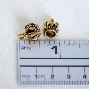 May include: Two gold-toned metal crown-shaped bead caps with intricate details. The caps are approximately 1 cm in diameter and have a small loop on one side. The caps are shown next to a ruler for size reference.