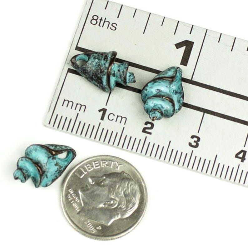 May include: Three turquoise-colored seashell beads with a distressed finish, displayed with a ruler showing measurements in centimeters and a US dime for size comparison. The beads have a spiral design and are made of metal.