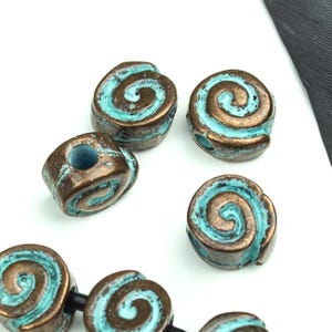 May include: A collection of round beads featuring a spiral design. Each bead has a metallic bronze finish with a weathered turquoise patina in the recessed areas. The beads are shaped like small coils and include a central hole for stringing.