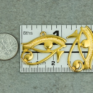 May include: Two gold-colored metal charms in the shape of the Eye of Horus, a symbol of protection in ancient Egyptian culture. The charms are about 2 inches long and are shown next to a ruler for size reference.