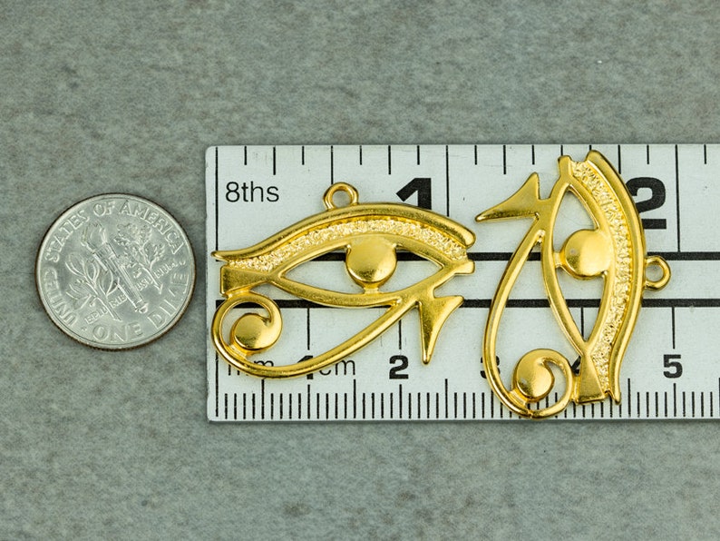 May include: Two gold-colored metal charms in the shape of the Eye of Horus, a symbol of protection in ancient Egyptian culture. The charms are about 2 inches long and are shown next to a ruler for size reference.