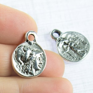 May include: Two silver-toned metal charms in the shape of a round coin with a raised design of a woman's head wearing a helmet. The charms have a small loop at the top for attaching to a necklace or bracelet.