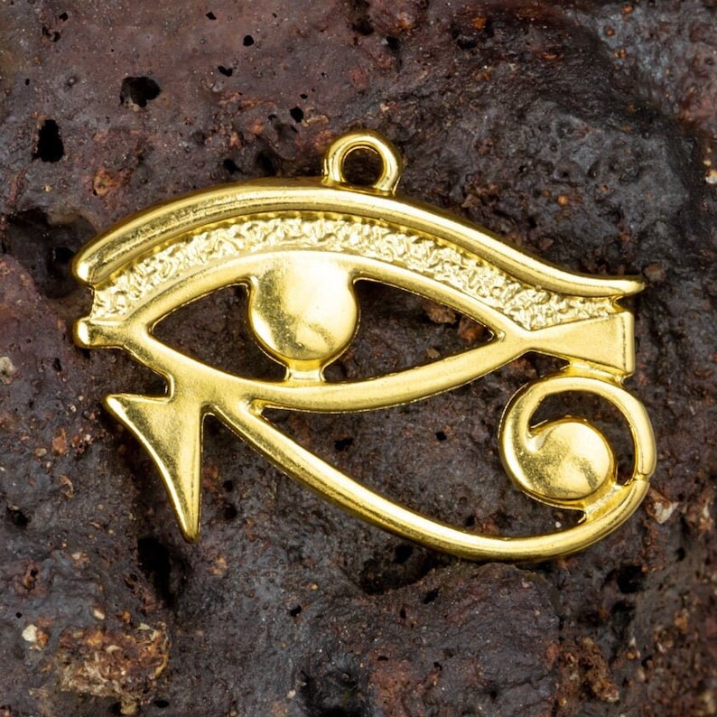 May include: A gold-colored metal charm in the shape of the Eye of Horus, an ancient Egyptian symbol of protection and good health. The charm has a loop at the top for attaching to a necklace or bracelet.