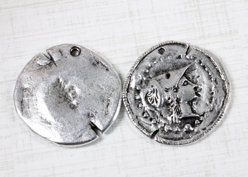 K&ouml;nnte beinhalten: Zwei silberfarbene Metall-M&uuml;nzenanh&auml;nger mit einem Loch zum Aufh&auml;ngen. Ein Anh&auml;nger zeigt ein detailliertes Design eines Frauenprofils, w&auml;hrend der andere Anh&auml;nger schlicht ist.