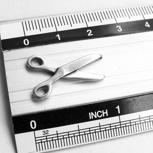 May include: A silver pair of scissors measuring approximately 1 inch in length, placed on a ruler with inch markings.