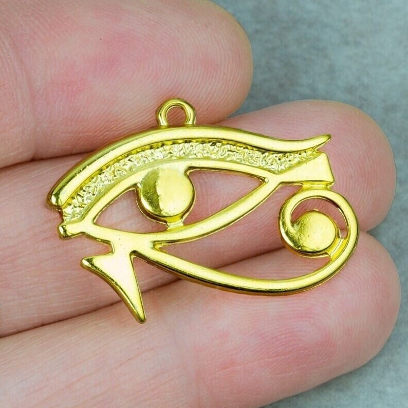 May include: Gold-colored metal pendant charm in the shape of the Eye of Horus, an ancient Egyptian symbol of protection and good health.