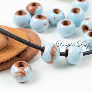 May include: A close-up of a necklace with light blue and brown ceramic beads. The beads have a speckled pattern and are strung on a black cord. The necklace is lying on a brown surface. "Loopin Loop" is written in white text on the surface.