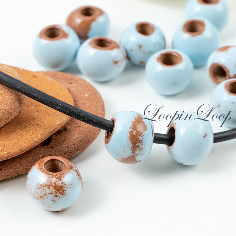 May include: A close-up of a necklace with light blue and brown ceramic beads. The beads have a speckled pattern and are strung on a black cord. The necklace is lying on a brown surface. "Loopin Loop" is written in white text on the surface.