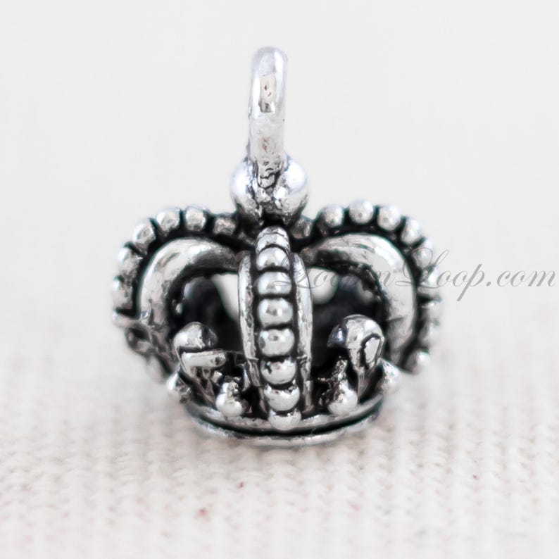 May include: Silver crown charm with a detailed design. The crown has a pointed top and a rounded base.