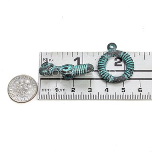 May include: Two antique-style metal charms, one shaped like a ring and the other like a stylized figure, both with a turquoise patina. The charms are measured against a ruler, with the ring charm measuring approximately 2 cm in diameter.