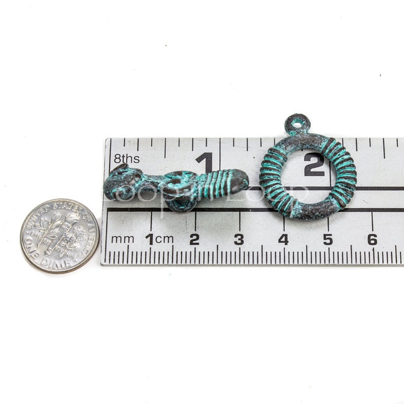 May include: Two antique-style metal charms, one shaped like a ring and the other like a stylized figure, both with a turquoise patina. The charms are measured against a ruler, with the ring charm measuring approximately 2 cm in diameter.