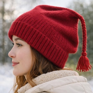May include: A knitted red hat with a long, braided tassel. The hat is a deep red color and has a ribbed brim. The tassel ends in a small, decorative knot. The hat is worn by a person.