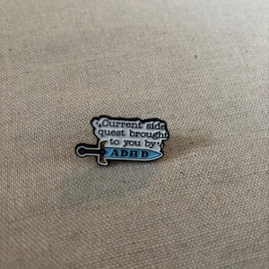 May include: A small enamel pin with a sword design. The pin has the text "Current side quest brought to you by ADHD" in a speech bubble. The sword has a blue blade and gold hilt.