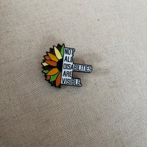 Peut inclure: Épingle en émail en forme de tournesol avec une bordure noire. Le tournesol a des pétales jaunes, oranges et bruns. L'épingle indique "NOT ALL DISABILITIES ARE VISIBLE" en texte noir.