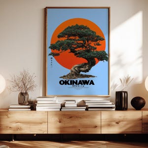 May include: Framed art print featuring a bonsai tree in front of a large orange sun against a light blue background. The word "OKINAWA" is displayed in white text at the bottom. The print is in a wooden frame.