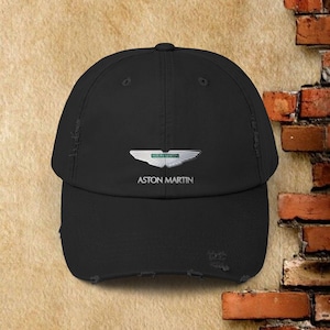 May include: Black baseball cap with a distressed brim. The cap features the Aston Martin logo in silver and green, with the brand name printed below. The cap is made of a durable fabric and has a curved peak.