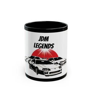 May include: A white ceramic mug with a black rim and interior. The mug features the text "JDM LEGENDS" above a red sun graphic and illustrations of several sports cars. The cars are in black and white.