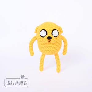 Könnte beinhalten: Ein gelber, gehäkelter Stoffhund mit schwarzen Augen und einer schwarzen Nase. Der Hund steht auf einem weißen Hintergrund.