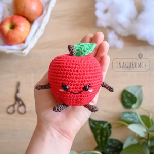 May include: A handmade red crochet apple with a smiling face, green leaf, and brown stem and limbs. The apple is held in a hand. The text "ENAGURUMIS" is in the background.