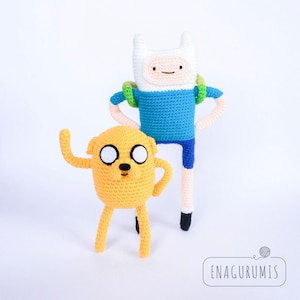 May include: Two handmade crochet figures of cartoon characters, Finn and Jake, from Adventure Time. Finn is tall with a white hat, blue shirt, and backpack. Jake is a yellow dog-like character. The figures are on a white background.