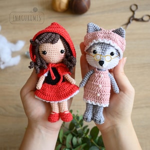 May include: Two handmade crochet dolls: Little Red Riding Hood in a red dress and cape, and a wolf dressed as a grandmother in a pink dress and hat. The dolls are held in hands, showcasing the detailed craftsmanship and whimsical design.