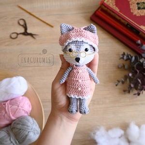 Op de afbeelding: Gehaakte wolfpop in een roze jurk, hoed en bril. De pop heeft grijs en wit haar. De afbeelding bevat een schaar en de tekst 'ENAGURUMIS'. Handgemaakt.