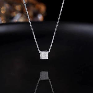 May include: A silver necklace featuring a small, textured cube pendant. The pendant hangs from a delicate chain, creating a minimalist and modern aesthetic. The necklace is displayed against a dark background, highlighting its metallic shine.