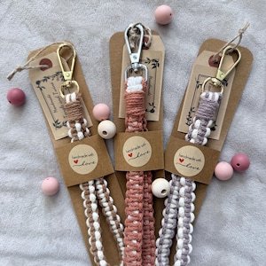 Puede incluir: Tres llaveros de macramé hechos a mano en marrón, rosa y morado con detalles blancos. Cada llavero tiene un cierre de metal y está unido a una etiqueta marrón con las palabras "handmade with love". Hay cuentas rosas y blancas esparcidas.