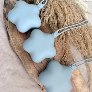 May include: Three light blue star-shaped silicone pacifier clips with attached loops. The clips are arranged on a wooden board with dried grasses in the background. The clips are designed to attach to a pacifier.