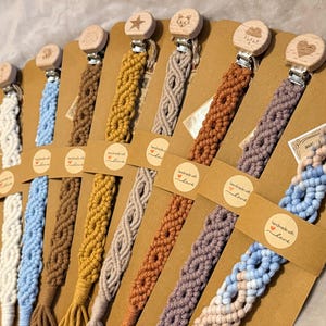 Op de afbeelding: Een verzameling macramé fopspeenclips in verschillende kleuren, waaronder blauw, wit, bruin en geel. Elke clip heeft een houten kraal met een ander ontwerp, zoals een ster, beer of hart. De clips worden gepresenteerd op bruin karton.