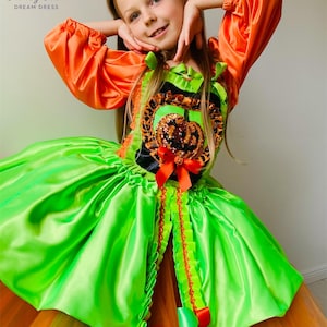 May include: A colourful Halloween costume with a pumpkin theme, featuring a green and orange dress. The dress has a satin bodice with a sequined pumpkin design, orange sleeves, and a layered skirt. Red bows and trim add detail.