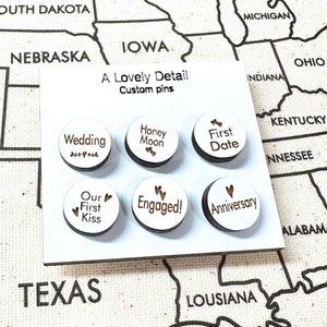 May include: A set of six round wooden pins with different phrases printed on them. The phrases are "Wedding", "Honey Moon", "First Date", "Our First Kiss", "Engaged!" and "Anniversary". The pins are on a white background with a map of the United States behind them.